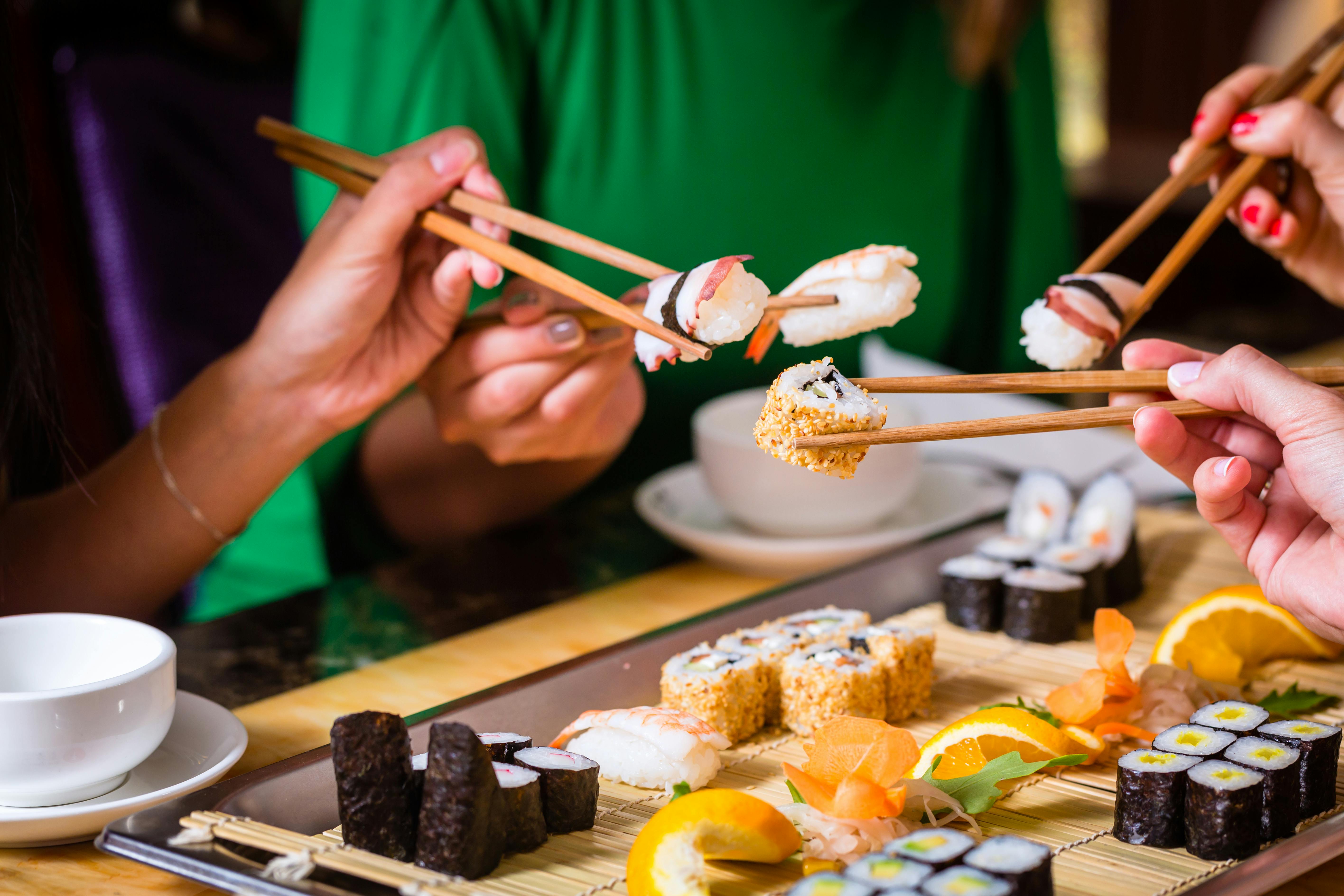 Three people use chopsticks to pick up pieces of sushi from a platter filled with various sushi rolls and garnishes. A cup and saucer are visible on the table, and one person wears a green shirt.