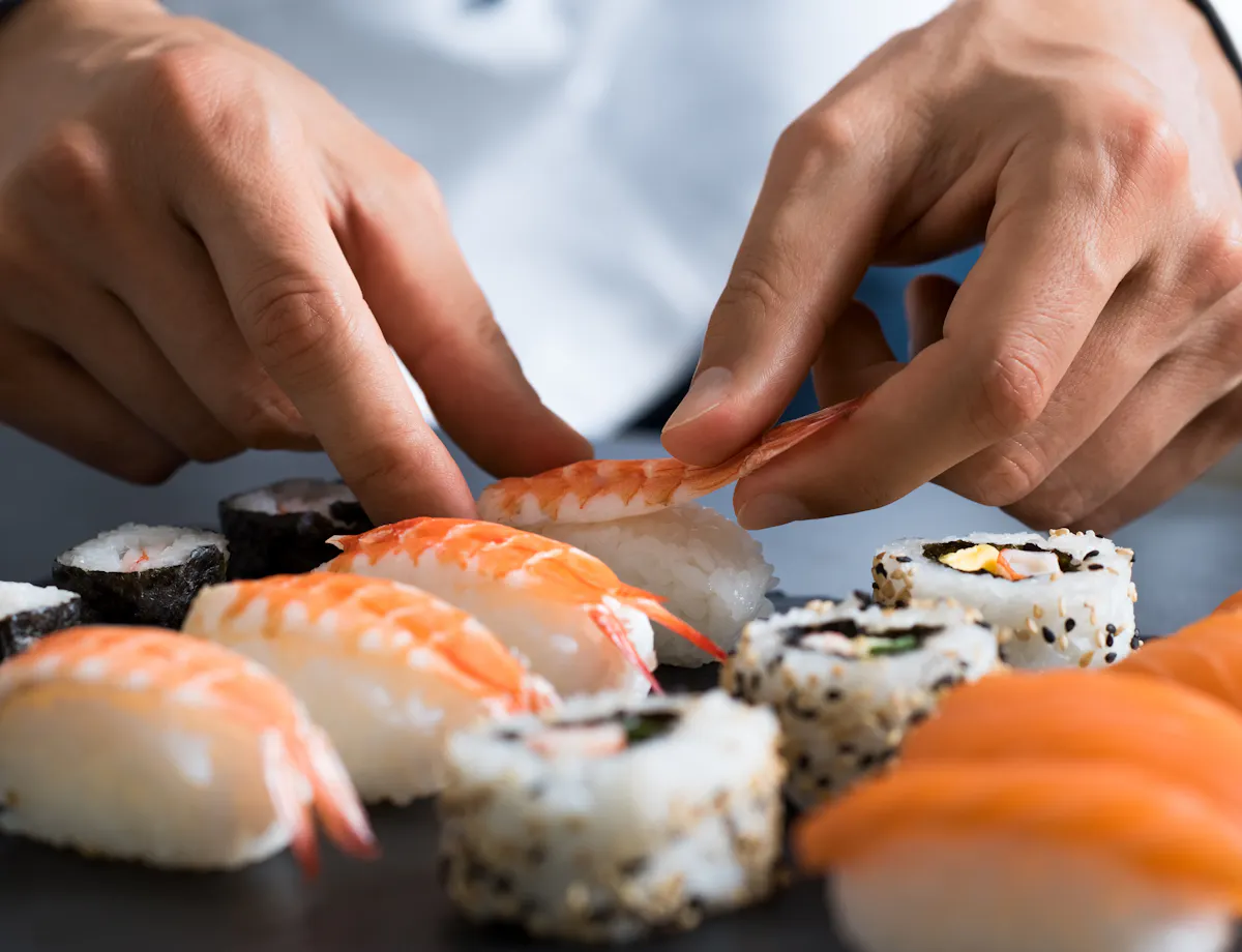Making Sushi Close-up of a chef’s hands carefully arranging pieces of shrimp nigiri and assorted sushi rolls on a dark surface.