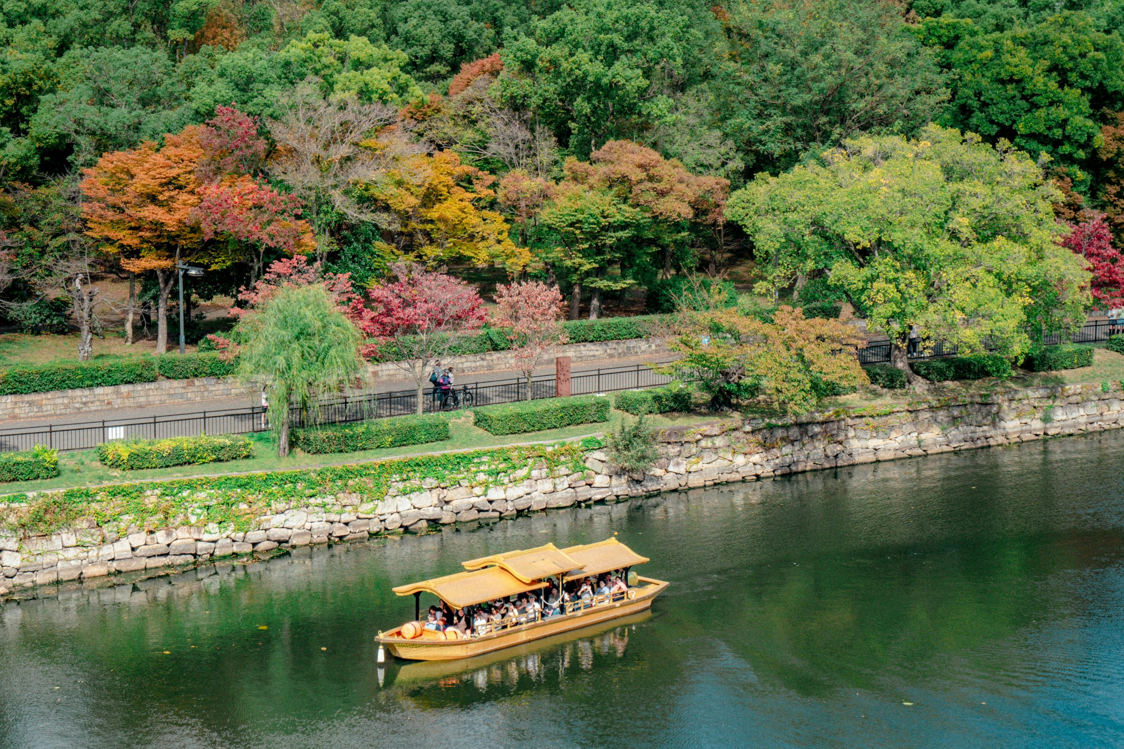 A traditional wooden boat with passengers glides along a calm river, bordered by a stone wall and vibrant autumn trees in a park.