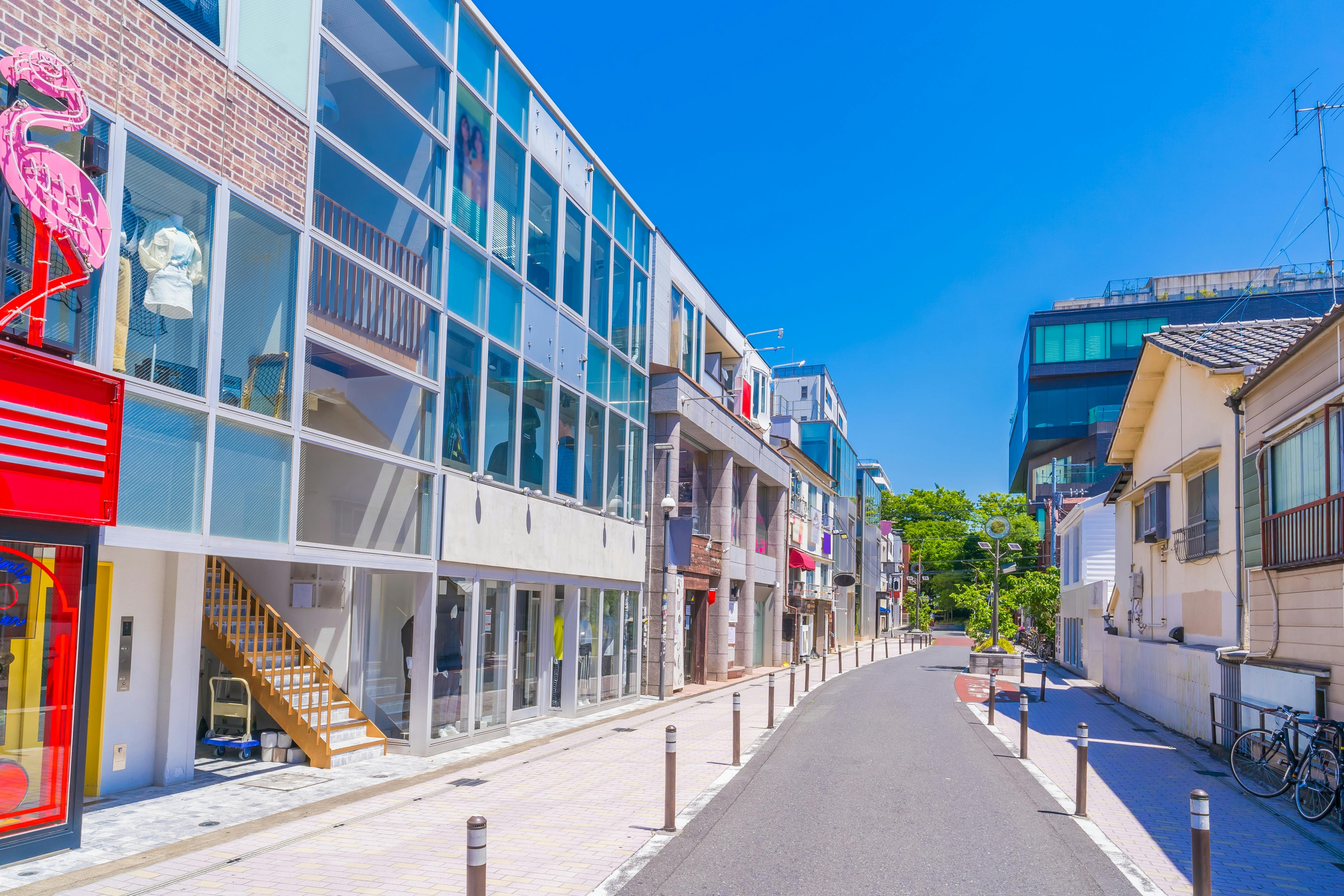 A quiet urban street with modern glass-fronted buildings, a red storefront featuring a pink flamingo sign, and traditional houses under a clear blue sky. No people are visible; the scene is bright and sunny.