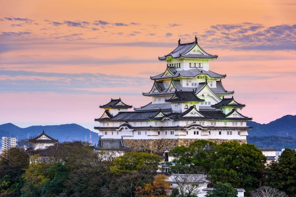 A white traditional Japanese castle with multiple tiered roofs stands on a stone base amid trees, set against a colorful sunset sky with distant mountains in the background.