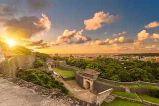 A view of Shuri Castle in Okinawa, Japan, at sunset. The sun glows behind lush green trees and ancient stone walls, with dramatic clouds and a cityscape in the background under a colorful sky.
