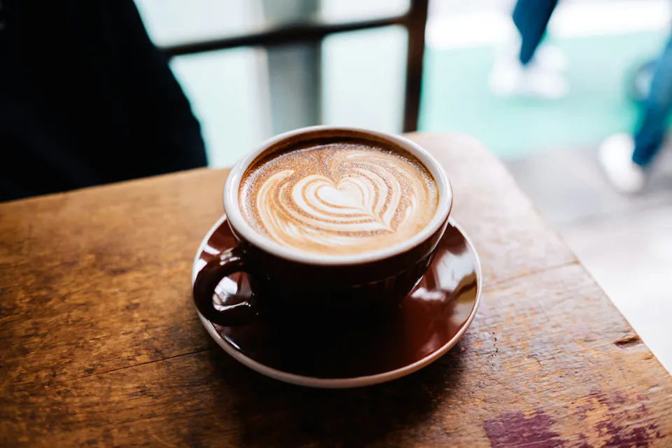 A cup of coffee with a heart-shaped latte art design on top, sitting on a brown saucer on a wooden table. The focus is on the coffee, with a softly blurred background indicating the interior of a cafe.