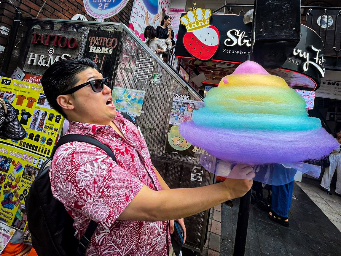A man wearing sunglasses and a pink patterned shirt looks surprised as he holds a large, colorful, swirled cotton candy on a busy street lined with shops and signs.