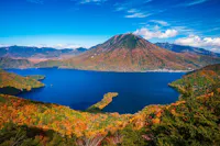 A deep blue lake surrounded by vibrant autumn foliage, with a tall, cone-shaped mountain in the background under a bright blue sky with scattered clouds.