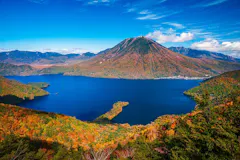 A deep blue lake surrounded by vibrant autumn foliage, with a tall, cone-shaped mountain in the background under a bright blue sky with scattered clouds.