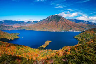 A deep blue lake surrounded by vibrant autumn foliage, with a tall, cone-shaped mountain in the background under a bright blue sky with scattered clouds.