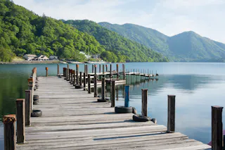 A wooden dock with black tires along the sides extends over a calm lake, surrounded by green forested hills and mountains under a bright blue sky.