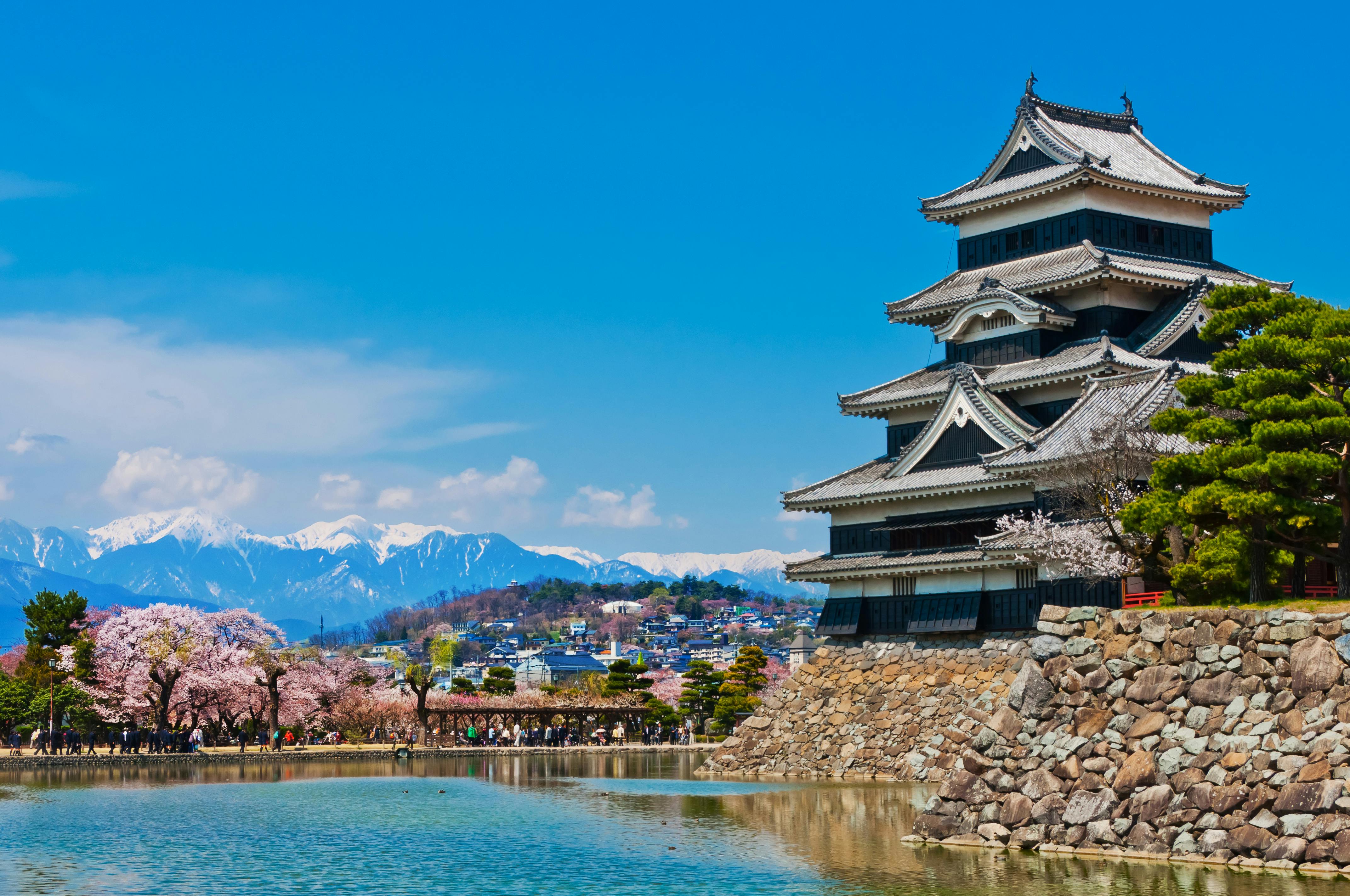 A traditional Japanese castle stands beside a calm moat, with cherry blossom trees in bloom and snow-capped mountains in the background under a clear blue sky.