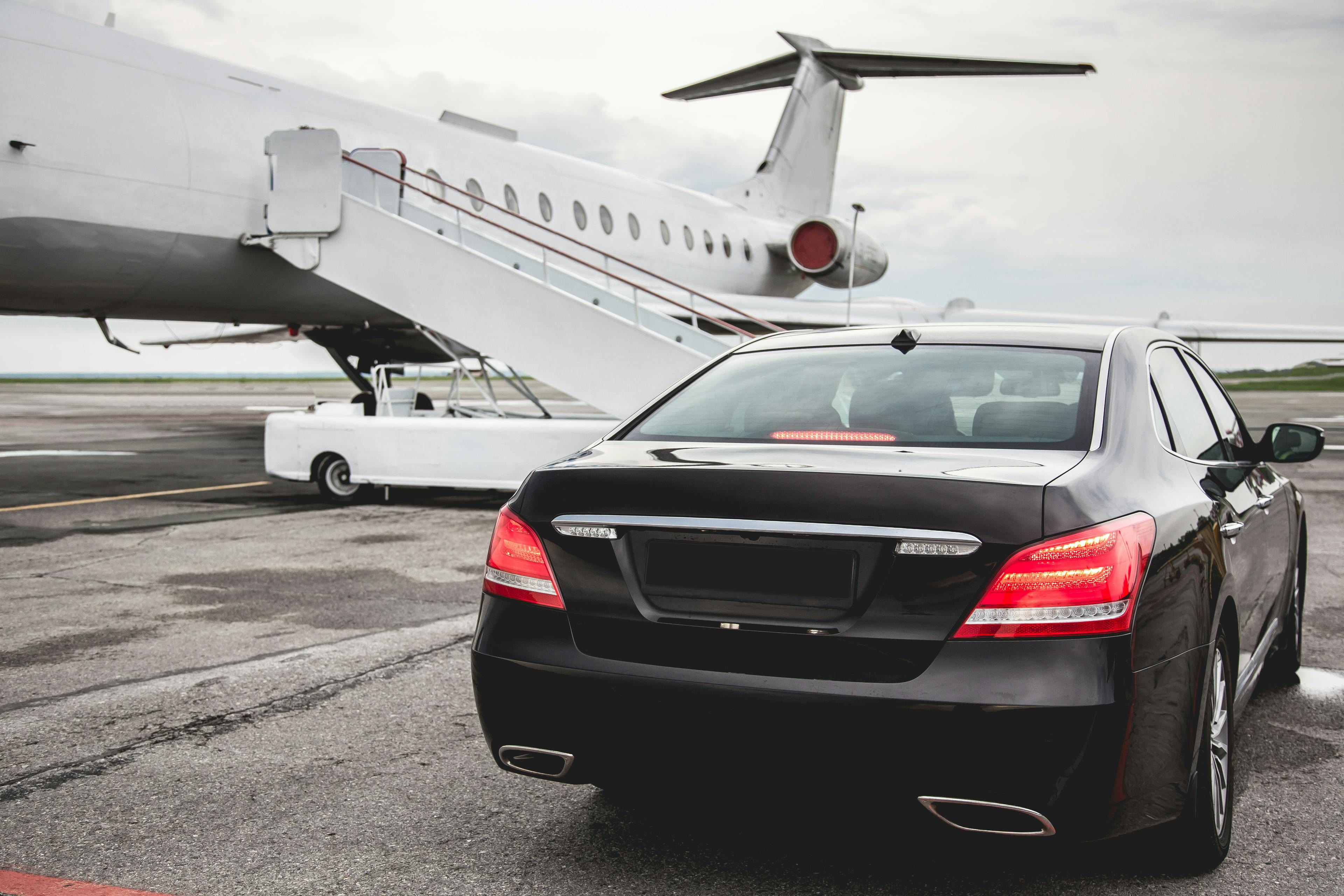 A black luxury car is parked on an airport tarmac near a private jet with an airstair. The scene suggests a setting of luxury travel and exclusivity.
