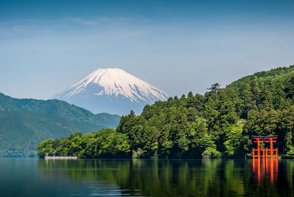 A tranquil scene of Mount Fuji with a snow-capped peak, surrounded by a dense forest. A red torii gate stands by the edge of a calm lake, reflecting the vibrant greenery and clear blue sky.