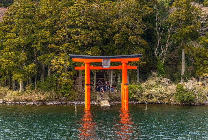 A large red torii gate stands in the water near the shore, with a stone staircase leading into a dense forest in the background. Several people are visible near the stairs and gate.