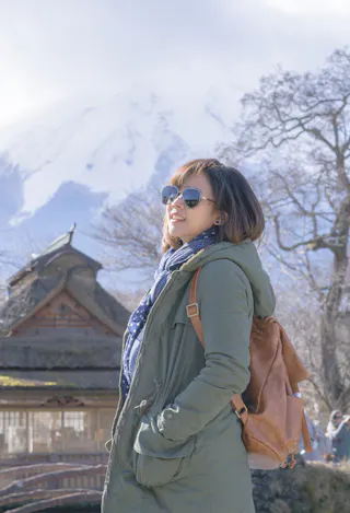 A woman wearing sunglasses, a green coat, and a brown backpack stands outdoors, smiling. Behind her are bare trees, a traditional wooden building, and a snow-covered mountain in the background.