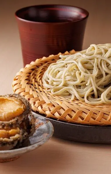 Soba A serving of cold soba noodles on a bamboo tray, with sliced abalone on a small plate and a wooden cup in the background, set on a light-colored table.