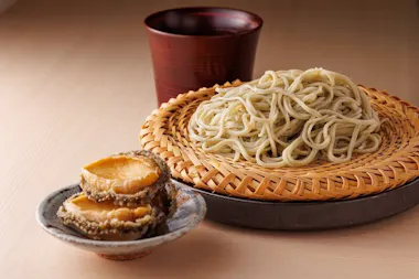 A serving of cold soba noodles on a bamboo tray, with sliced abalone on a small plate and a wooden cup in the background, set on a light-colored table.
