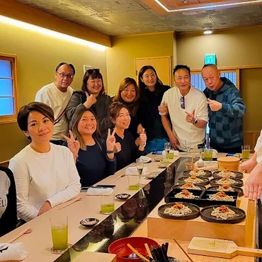 Soba Making A group of people pose and smile around a sushi bar inside a restaurant, with green drinks and plates of food on the counter. A chef in a white uniform stands at the end, also smiling.