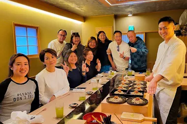 A group of people pose and smile around a sushi bar inside a restaurant, with green drinks and plates of food on the counter. A chef in a white uniform stands at the end, also smiling.