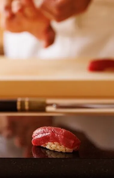 Sushi A piece of sushi with red fish sits on a dark counter. In the background, a chef shapes another piece of sushi near a cutting board and a knife, with their hands slightly out of focus.