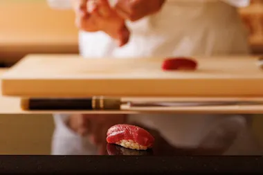 A piece of sushi with red fish sits on a dark counter. In the background, a chef shapes another piece of sushi near a cutting board and a knife, with their hands slightly out of focus.