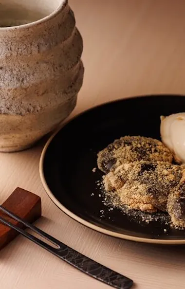 Sushi A black plate with mochi covered in kinako powder and a scoop of ice cream, next to a rustic ceramic mug, a small cup, and metal chopsticks resting on a wooden holder.