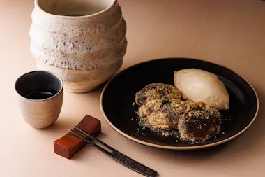 A black plate with mochi covered in kinako powder and a scoop of ice cream, next to a rustic ceramic mug, a small cup, and metal chopsticks resting on a wooden holder.