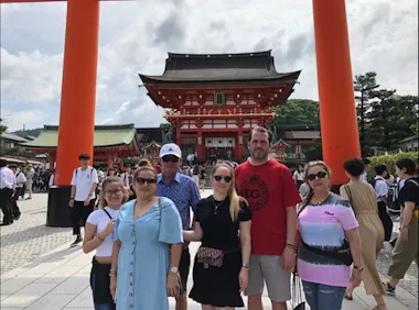 A group of six people pose for a photo in front of a large red torii gate at a busy Japanese shrine, with traditional buildings and visitors visible in the background on a sunny day.