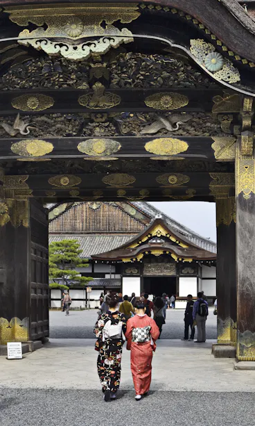 Nijo Castle Two women in traditional kimonos walk under an ornate wooden gate with gold details at a historic Japanese site, with more visitors and a large building visible in the background.