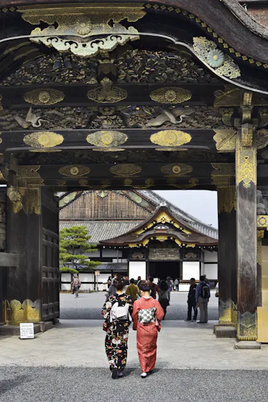 Two women in traditional kimonos walk under an ornate wooden gate with gold details at a historic Japanese site, with more visitors and a large building visible in the background.