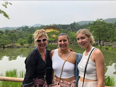 Three smiling women pose in front of a pond with lush greenery. In the background stands a golden building, likely Kinkaku-ji in Japan, surrounded by trees and mountains on a cloudy day.
