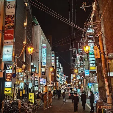 Hiroshima Food Tour A lively city street at night with people walking, illuminated by streetlights and colorful signs on tall buildings. Bicycles are parked on the left, and cars drive down the road under a tangle of overhead wires.
