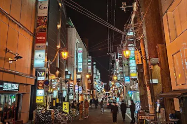 A lively city street at night with people walking, illuminated by streetlights and colorful signs on tall buildings. Bicycles are parked on the left, and cars drive down the road under a tangle of overhead wires.