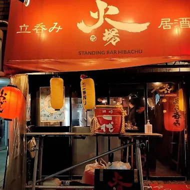 Hiroshima Food Tour Street view of a Japanese standing bar called "Standing Bar Hibachu," with a red awning, illuminated red and yellow lanterns, and a person working behind the counter inside.