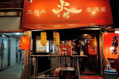 Street view of a Japanese standing bar called "Standing Bar Hibachu," with a red awning, illuminated red and yellow lanterns, and a person working behind the counter inside.