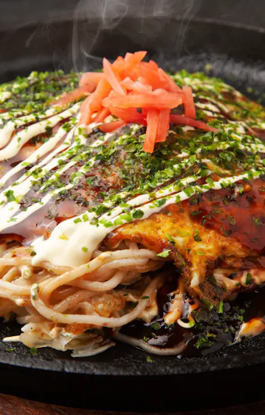 Okonomiyaki A hot plate of okonomiyaki topped with mayonnaise, dark sauce, chopped green herbs, and pickled ginger sits steaming on a black skillet, with noodles and vegetables visible underneath.