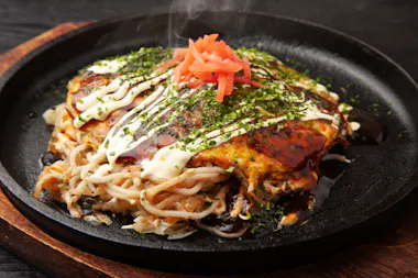 A hot plate of okonomiyaki topped with mayonnaise, dark sauce, chopped green herbs, and pickled ginger sits steaming on a black skillet, with noodles and vegetables visible underneath.