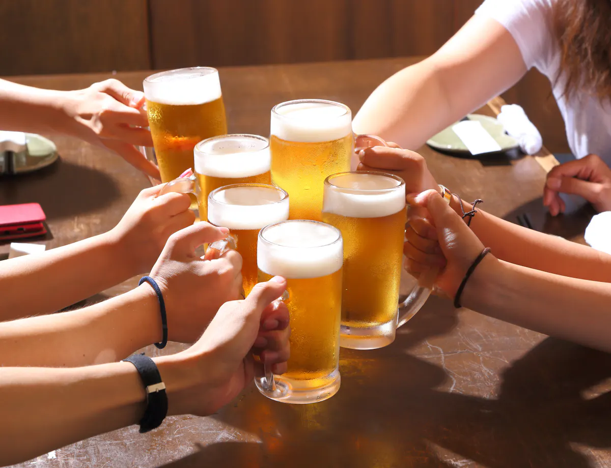 Toast Several people raise glasses of beer together over a wooden table, toasting in a celebratory gesture. The scene is bright and cheerful, suggesting a social gathering or celebration.