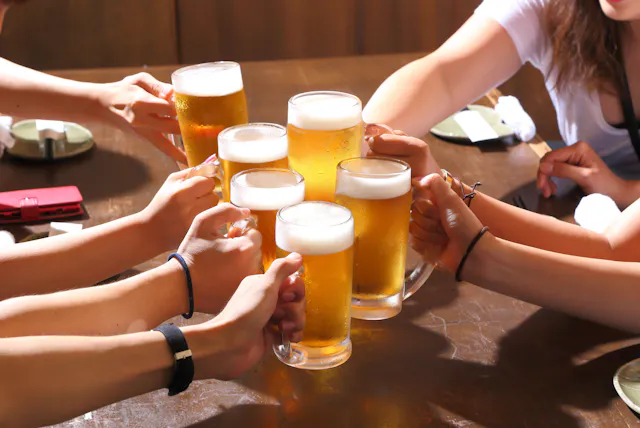 Several people raise glasses of beer together over a wooden table, toasting in a celebratory gesture. The scene is bright and cheerful, suggesting a social gathering or celebration.