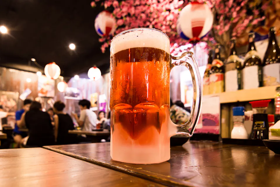 A frosty mug of beer sits on a wooden table in a cozy, dimly-lit bar. The background features blurred patrons and Japanese decor, including hanging lanterns and cherry blossom decorations, creating a warm and inviting atmosphere.