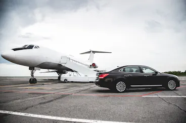 A black luxury car is parked on an airport tarmac next to a white private jet with stairs extended, under a cloudy sky.