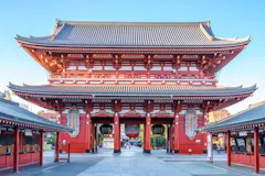 The main gate of Senso-ji Temple in Tokyo, Japan, featuring a large red wooden structure with traditional curved roof, lanterns, and intricate decorative details, seen against a clear blue sky.