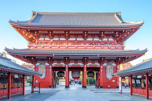 The main gate of Senso-ji Temple in Tokyo, Japan, featuring a large red wooden structure with traditional curved roof, lanterns, and intricate decorative details, seen against a clear blue sky.