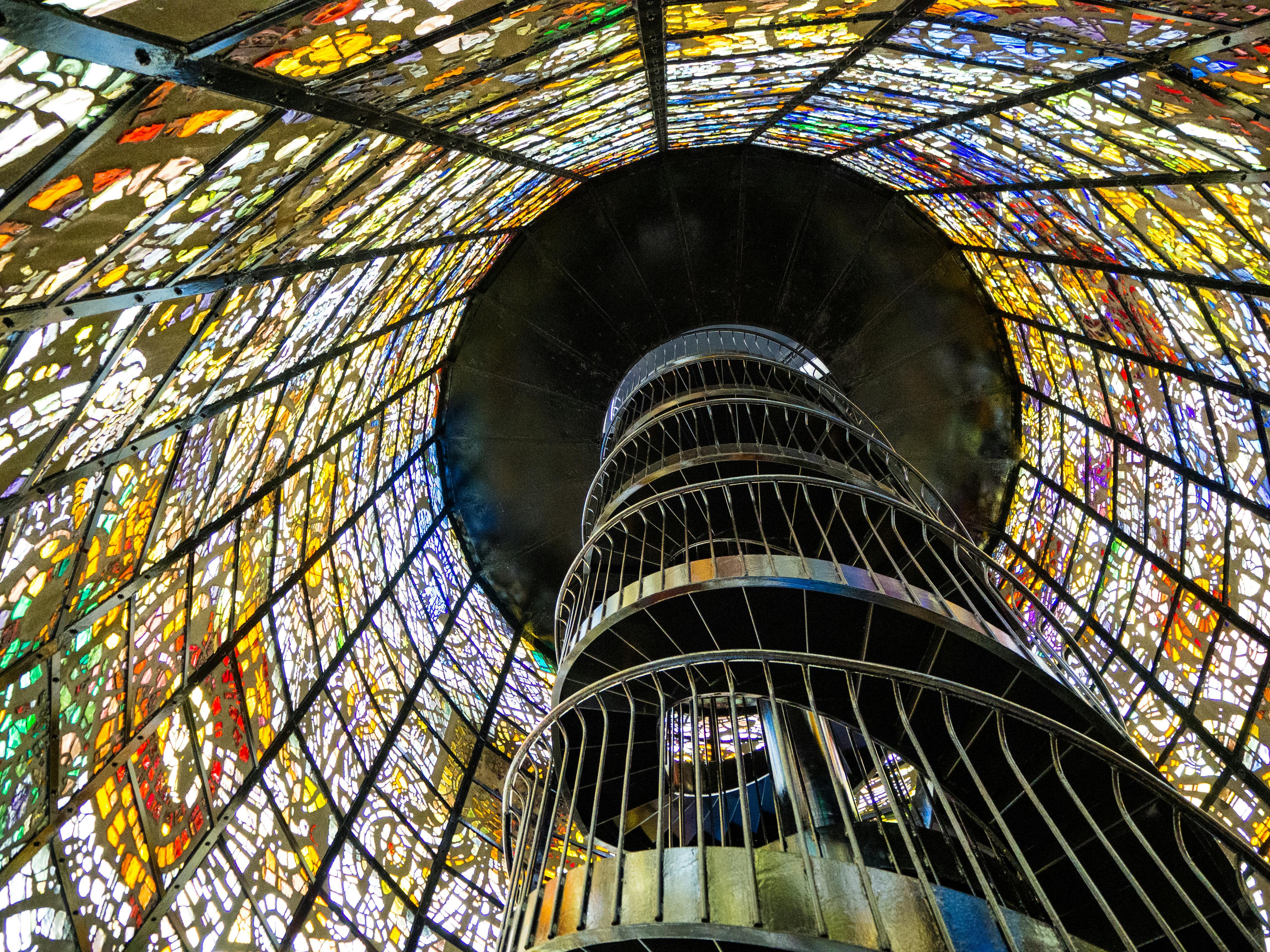 A spiral staircase rises inside a tall, cylindrical tower with colorful stained glass walls and ceiling, creating a vibrant, kaleidoscopic effect as light shines through the intricate glass patterns.