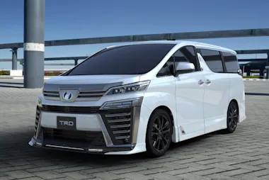 A white, modern minivan with a sleek, angular design, black wheels, tinted windows, and a TRD badge is parked on a paved area under a clear blue sky.