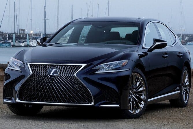 A black Lexus sedan is parked outdoors near a marina, featuring a large spindle grille and sleek, modern headlights with sailboats and water visible in the background.