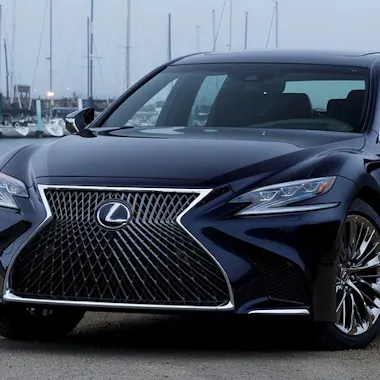 Sedan A black Lexus sedan is parked outdoors near a marina, featuring a large spindle grille and sleek, modern headlights with sailboats and water visible in the background.