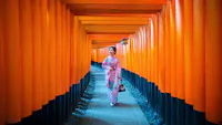 A woman in a pink kimono walks through a pathway lined with vibrant orange torii gates, likely at the Fushimi Inari Shrine in Kyoto, Japan. She is holding a small bag and looking to the side.