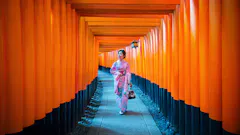 A woman in a pink kimono walks through a pathway lined with vibrant orange torii gates, likely at the Fushimi Inari Shrine in Kyoto, Japan. She is holding a small bag and looking to the side.