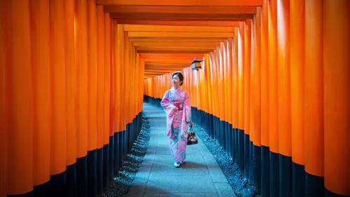 A woman in a pink kimono walks through a pathway lined with vibrant orange torii gates, likely at the Fushimi Inari Shrine in Kyoto, Japan. She is holding a small bag and looking to the side.