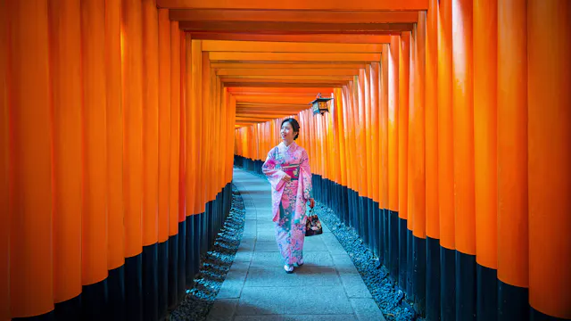 A woman in a pink kimono walks through a pathway lined with vibrant orange torii gates, likely at the Fushimi Inari Shrine in Kyoto, Japan. She is holding a small bag and looking to the side.