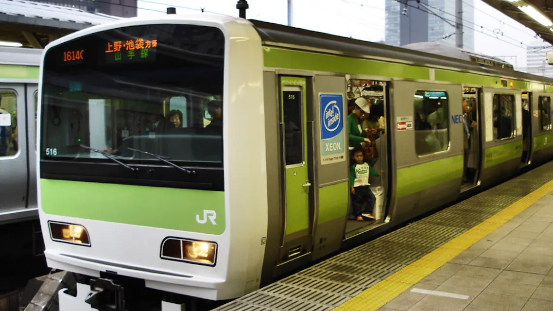 A green JR train is stopped at a platform with open doors. Passengers are entering and exiting. The platform is visible in the foreground, displaying yellow safety markers. The train's destination is shown in Japanese on the sign above.
